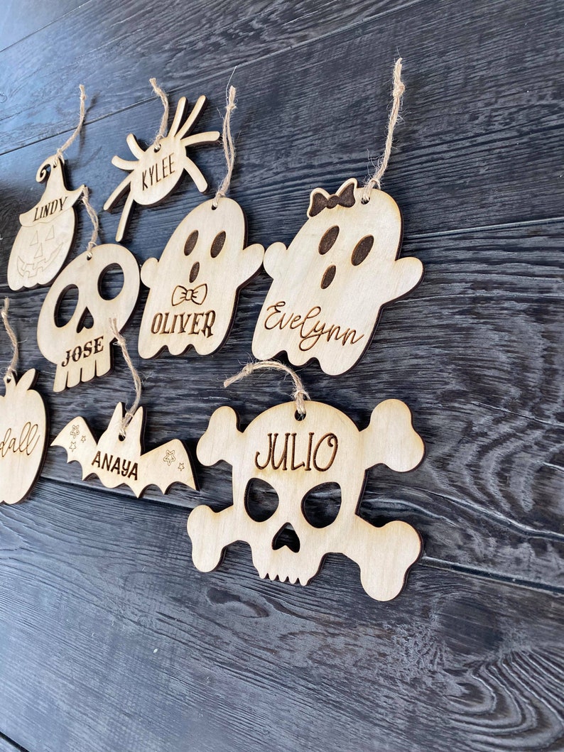 May include: A set of eight wooden Halloween ornaments with names written on them. The ornaments are shaped like a pumpkin, spider, ghost, skull, bat, and apple.