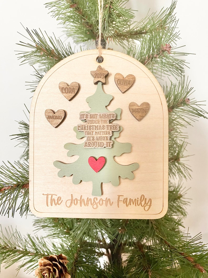 Peut inclure: Une d&eacute;coration de No&euml;l en bois en forme d'arbre avec le texte "Ce n'est pas ce qui se trouve sous le sapin de No&euml;l qui compte, mais qui est autour." L'ornement porte &eacute;galement les noms "Antonio", "Cora", "Olivia", "Martin" et "The Johnson Family".