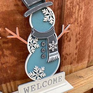 Puede incluir: Una figura de mu&ntilde;eco de nieve de madera con un sombrero de copa negro, un cuerpo azul y copos de nieve blancos. El mu&ntilde;eco de nieve sostiene un palo con una rama marr&oacute;n a cada lado. El mu&ntilde;eco de nieve est&aacute; de pie sobre una base blanca con la palabra "WELCOME" escrita en ella.