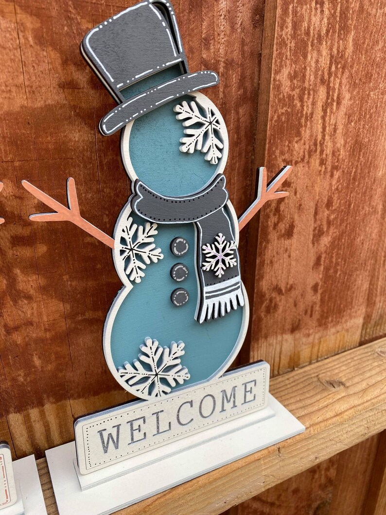 Puede incluir: Una figura de mu&ntilde;eco de nieve de madera con un sombrero de copa negro, un cuerpo azul y copos de nieve blancos. El mu&ntilde;eco de nieve sostiene un palo con una rama marr&oacute;n a cada lado. El mu&ntilde;eco de nieve est&aacute; de pie sobre una base blanca con la palabra "WELCOME" escrita en ella.