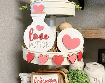 May include: A tiered tray decorated for Valentine's Day with a love potion sign, a heart-shaped sign, a sign that says "Love Lives Here", a sign that says "February 14", and a heart-shaped garland.