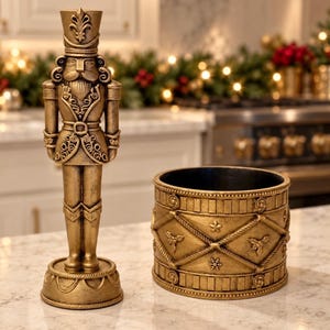 May include: A gold-colored nutcracker figurine standing on a circular base next to a matching gold-colored cylindrical container featuring a textured diamond pattern with embossed star, snowflake, and holly leaf details.