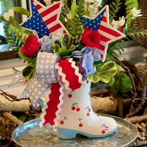 May include: A patriotic floral arrangement in a white boot-shaped vase with red cherry designs. The arrangement includes red, white, and blue flowers, star-shaped American flags, and a gingham ribbon. The vase sits on a metal tray.