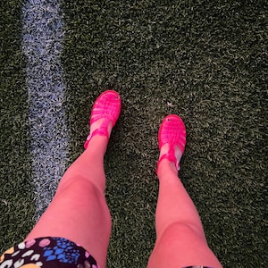 May include: A person wearing pink jelly sandals and a colorful patterned skirt standing on a green artificial grass surface.