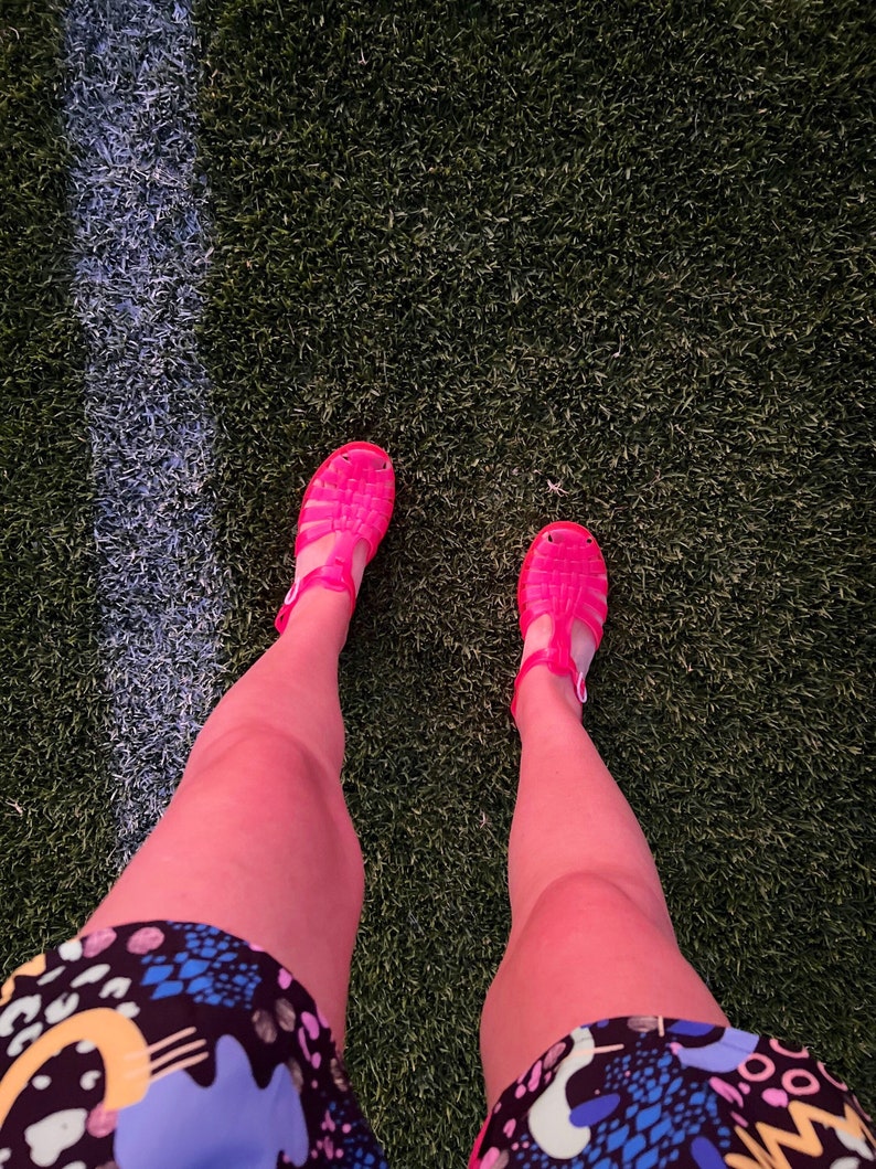 May include: A person wearing pink jelly sandals and a colorful patterned skirt standing on a green artificial grass surface.