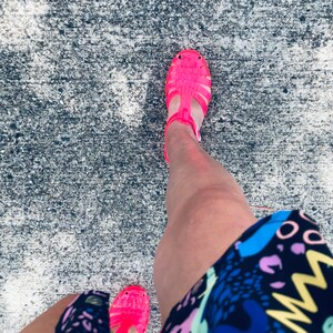 May include: A person wearing a black and white patterned skirt and bright pink jelly sandals. The sandals have a strap across the foot and an ankle strap.