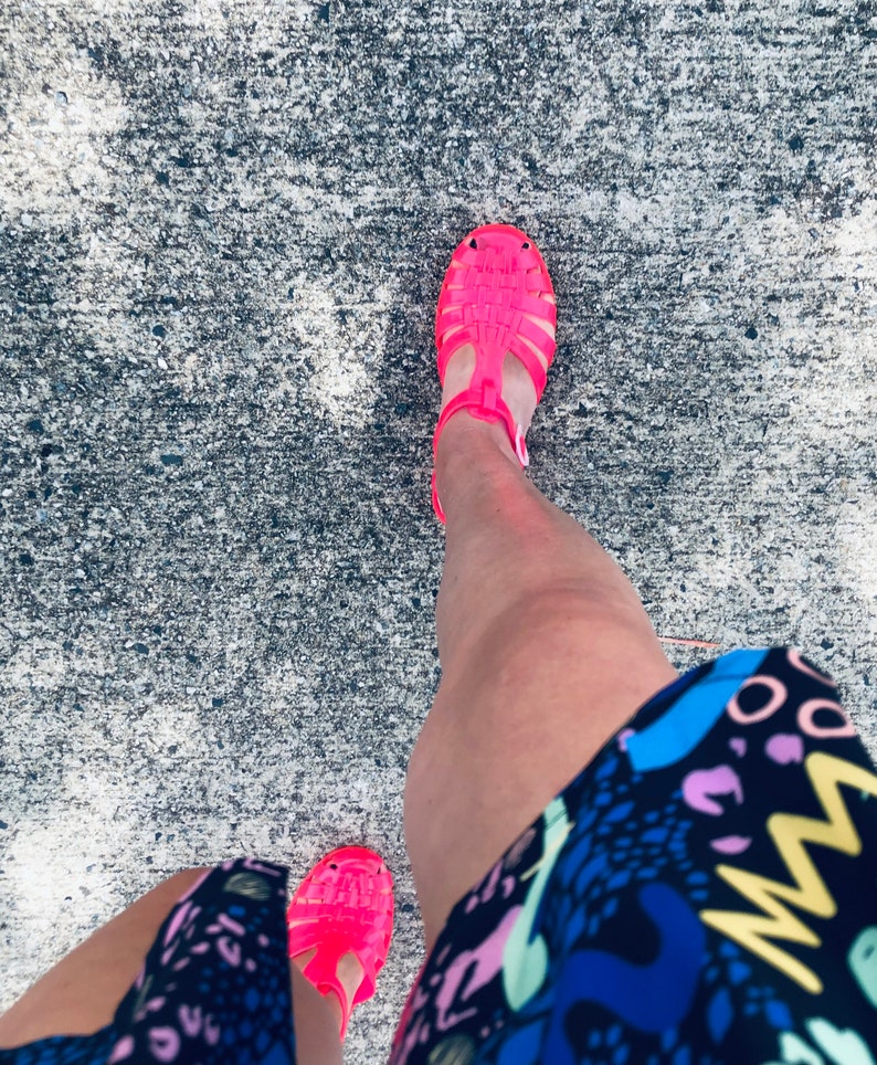 May include: A person wearing a black and white patterned skirt and bright pink jelly sandals. The sandals have a strap across the foot and an ankle strap.