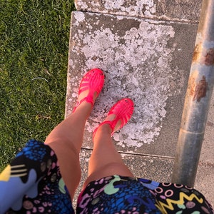 May include: A pair of bright pink jelly sandals with a strap across the foot and an open toe. The sandals are worn by a person wearing a colorful, patterned dress.