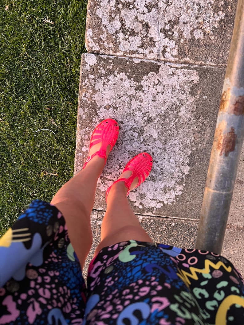 May include: A pair of bright pink jelly sandals with a strap across the foot and an open toe. The sandals are worn by a person wearing a colorful, patterned dress.