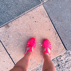 May include: A pair of bright pink jelly sandals with a criss-cross strap design. The sandals are worn by a person standing on a concrete surface.