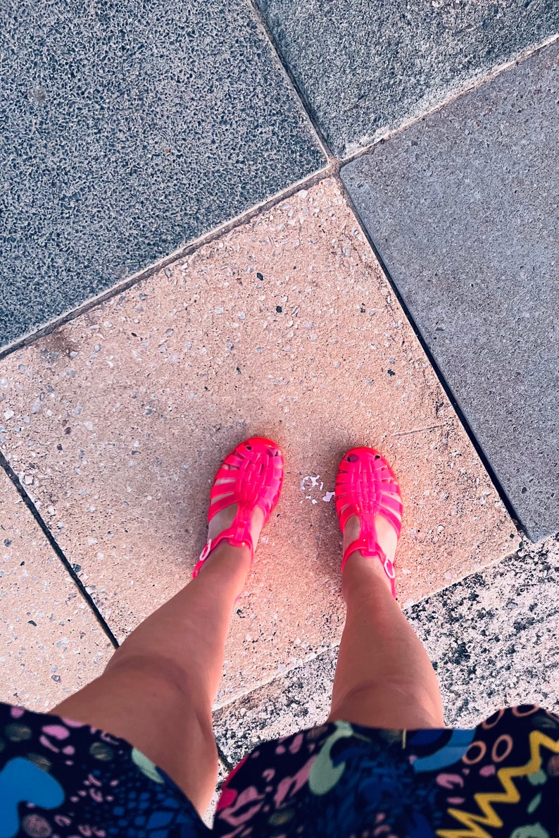 May include: A pair of bright pink jelly sandals with a criss-cross strap design. The sandals are worn by a person standing on a concrete surface.