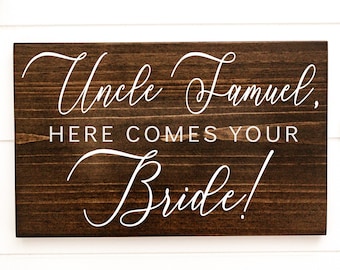 May include: A rectangular wooden sign with the words "Uncle Samuel, HERE COMES YOUR Bride!" in white script. The sign is stained a dark brown color and has a visible wood grain.