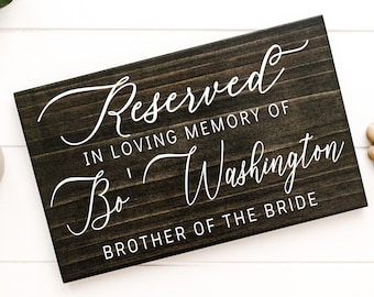 May include: A dark stained wooden sign with white cursive text that reads "Reserved in loving memory of Bo Washington, brother of the bride."