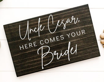 May include: A rectangular wooden sign with the text "Uncle Cesar, HERE COMES YOUR Bride!" in white script. The sign has a dark brown, rustic finish, and is likely used for a wedding.