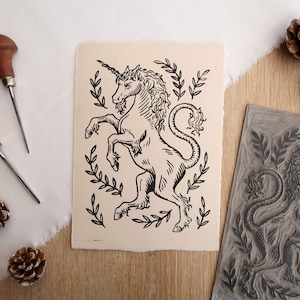 May include: A black and white linocut print of a unicorn with a leafy border. The print is on cream-coloured paper and is displayed next to a woodblock carving of the same design.