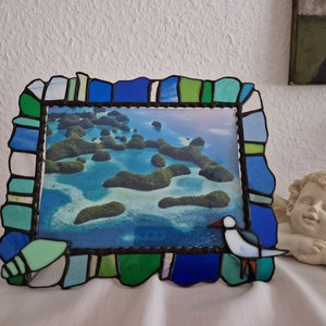 Può includere: Una cornice per foto in vetro colorato con un design a mosaico blu, verde e bianco. La cornice contiene una foto di un'isola tropicale con un uccello nell'angolo in basso a destra.