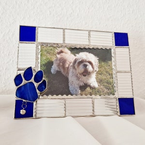 Könnte beinhalten: Ein blauer Pfotenabdruck aus Glasmalerei mit einem Glöckchen-Anhänger. Der Rahmen hat einen weißen Hintergrund und blaue Akzente. Ein Foto eines Hundes befindet sich im Rahmen.