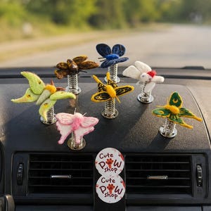 May include: A collection of colorful butterfly car air fresheners on a car dashboard. The butterflies are made of felt and sit on silver springs. Two circular air fresheners with the text "Cute Paw Shop" are also visible.