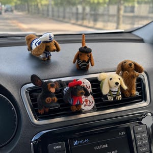 Può includere: Una collezione di figurine di cani bassotto fatte a mano, ognuna con abiti unici. Uno indossa un cappello da marinaio, un altro un berretto, e altri hanno camicie o cappotti a righe. Le figurine sono esposte sul cruscotto di un'auto.