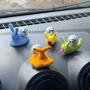 以下が含まれることがあります： 青、黄、緑、オレンジの4体のニードルフェルト製カタツムリのフィギュア。それぞれのカタツムリは、殻として小さな反射するミラーボールを背負っています。青と緑のカタツムリは舌を出しており、オレンジのカタツムリには2つの目があります。