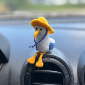 May include: A small, white felt bird wearing a yellow hat sits on a black car air vent. The bird has a blue scarf and yellow feet.