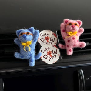 May include: Two felt cat air fresheners. One is blue with orange glasses and a yellow bow tie, the other is pink with pink polka dots, orange glasses, and a yellow bow. A circular tag with the text "Cute Paw Shop" is between them.