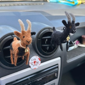 Könnte beinhalten: Zwei Filz-Ziegen-Lufterfrischer, die an den Lüftungsschlitzen eines Autos hängen. Die Ziege links ist braun und schwarz mit einer Glocke um den Hals. Die Ziege rechts ist schwarz mit einer Glocke um den Hals. Ein Aufkleber mit der Aufschrift "Cute Paw Shop" ist in der unteren rechten Ecke des Bildes zu sehen.