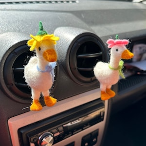 May include: Two felt goose car air fresheners. One goose wears a yellow flower hat and blue bow tie, the other wears a pink hat and green scarf. Both have orange feet and beaks. They are attached to the car's air vents.