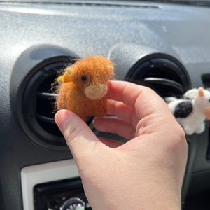 May include: A small, brown, fuzzy, felt toy cow with yellow ears is being held in a person's hand. The toy is in the interior of a car, near the air vent.