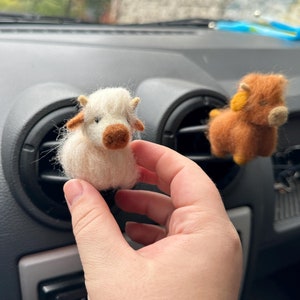 May include: Two small felt animal figurines, a white cow and a brown cow, are displayed in a car. The white cow is being held by a hand.
