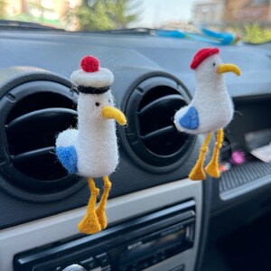 May include: Two felt seagull car ornaments, one wearing a red and white sailor hat, hanging from the air vents in a car. The seagulls are white with blue accents and yellow feet.