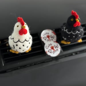 May include: Two felt chicken air fresheners, one white with black spots and the other black with white spots, both with red combs and yellow feet. The air fresheners are placed on a car vent. Two round white air freshener refills are also visible.