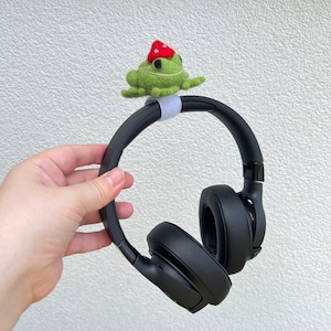 May include: Black over-ear headphones with a felt frog decoration. The frog is green with black eyes and a red mushroom hat. The headphones are held by a hand against a white wall.