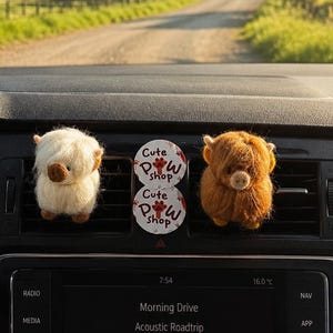 May include: Two fuzzy highland cow car air freshener clips, one white and one brown, attached to a vehicle dashboard vent. Between them are two circular white tags with the text Cute Paw Shop printed in red and black, featuring a paw print icon.