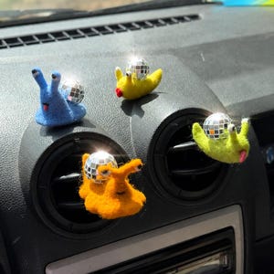 May include: Four felted snail figures in blue, yellow, and orange, each featuring a small reflective disco ball as a shell. The snails have distinct facial features and are positioned on the air vents and dashboard of a vehicle interior.