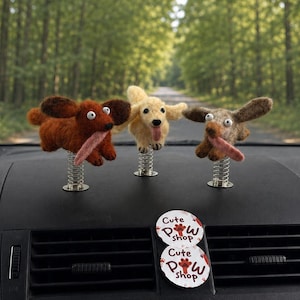 Może przedstawiać: Trzy figurki psów z filcu igłowego w kolorze brązowym, kremowym i szarym, każda zamontowana na metalowej sprężynie. Psy mają ruchome oczy i długie różowe języki. Dwie okrągłe białe naklejki z napisem "Cute Paw Shop" są przyklejone do nawiewu.