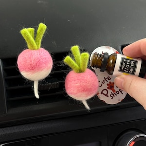 May include: Two pink and white felt radish car air fresheners with green tops. A hand is holding a small bottle of essential oil with the text "flora secret" on the label. A round sticker with the text "Cute P Shop" is also visible.