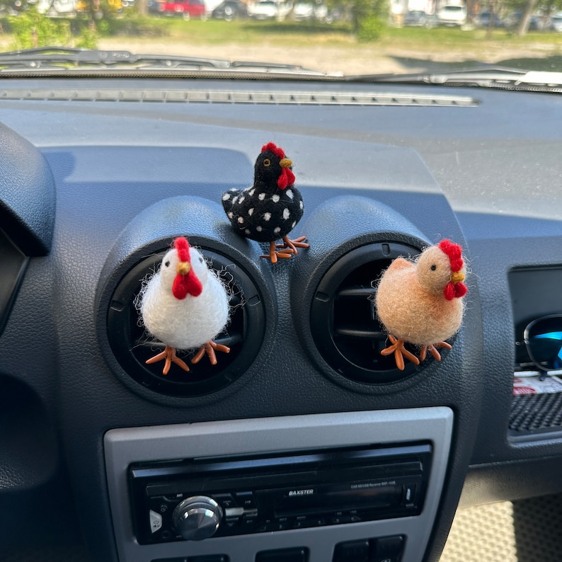 May include: Three felt chicken figurines hanging from the air vents of a car's dashboard. One chicken is black and white, one is white, and one is tan.