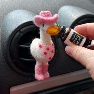 May include: A white felt goose wearing a pink cowboy hat and pink boots, with pink polka dots on its chest, is being used to diffuse essential oil from a small brown bottle labelled "flora secret".
