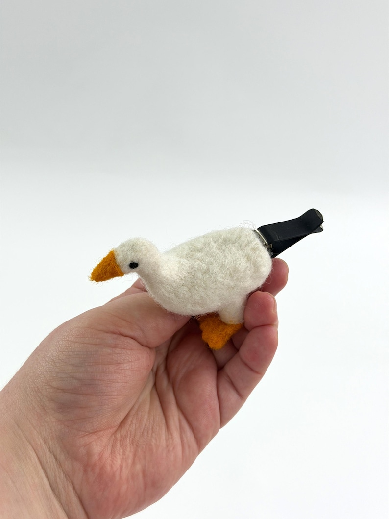 K&ouml;nnte beinhalten: Eine aus wei&szlig;er Wolle gefilzte Gans als Dekoration mit einem leuchtend orangefarbenen Schnabel und orangefarbenen F&uuml;&szlig;en. Die Gans hat ein kleines schwarzes Auge und ist an der R&uuml;ckseite mit einer schwarzen Metallklammer versehen.