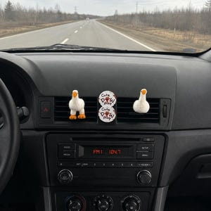 K&ouml;nnte beinhalten: Zwei wei&szlig;e, gefilzte G&auml;nse-L&uuml;ftungsclips mit orangefarbenen Schn&auml;beln und F&uuml;&szlig;en an einer Auto-L&uuml;ftungsd&uuml;se, platziert neben einer runden, wei&szlig;en Duftscheibe mit der Aufschrift Cute Paw Shop in schwarzer und roter Schrift.