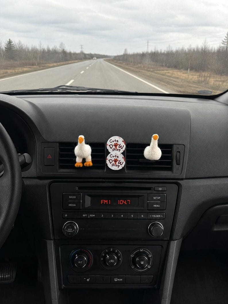 K&ouml;nnte beinhalten: Zwei wei&szlig;e, gefilzte G&auml;nse-L&uuml;ftungsclips mit orangefarbenen Schn&auml;beln und F&uuml;&szlig;en an einer Auto-L&uuml;ftungsd&uuml;se, platziert neben einer runden, wei&szlig;en Duftscheibe mit der Aufschrift Cute Paw Shop in schwarzer und roter Schrift.