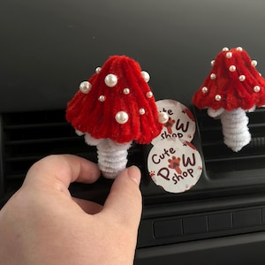 May include: Two red mushroom car air fresheners with white pearl embellishments. The mushroom caps are a bright red, and the stems are a fluffy white. A sticker with the text "Cute Paw Shop" is also visible.