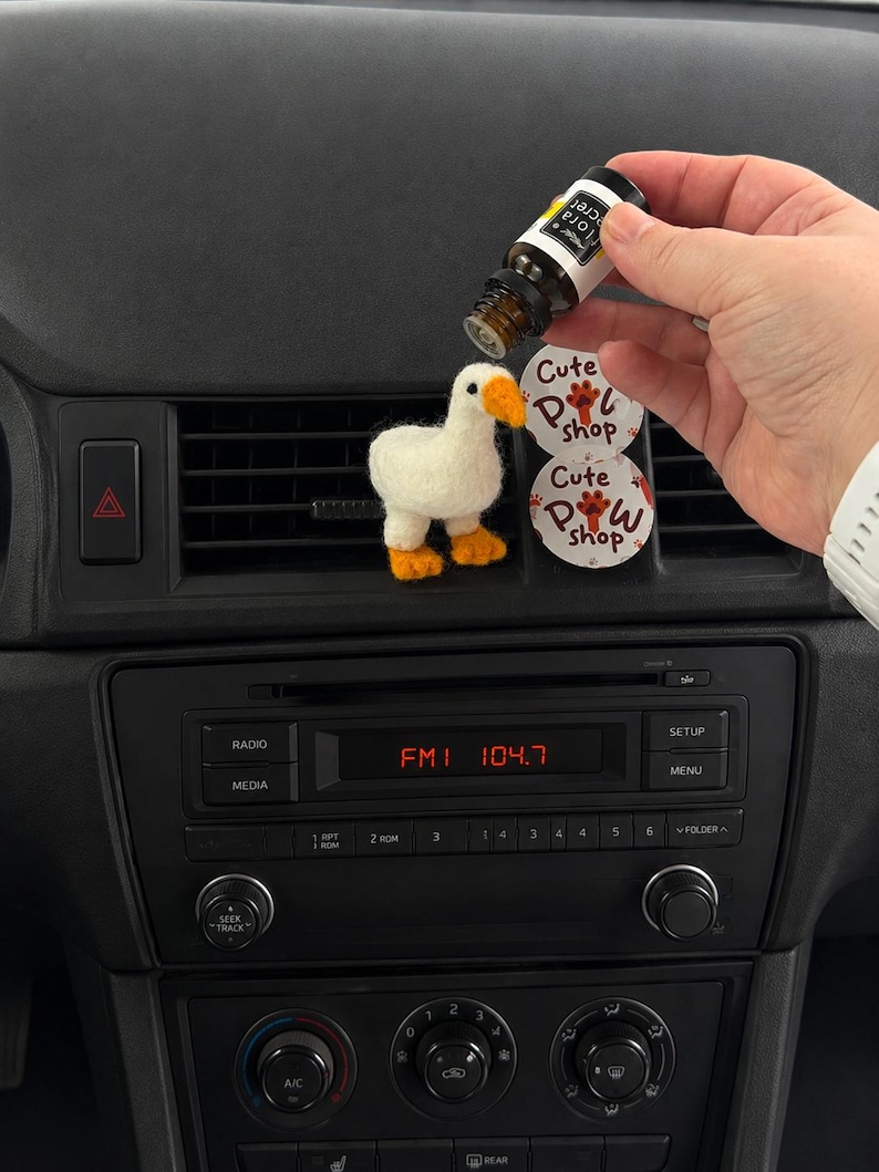 K&ouml;nnte beinhalten: Eine kleine, wei&szlig; nadelgefilzte G&auml;nsefigur mit orangefarbenem Schnabel und orangefarbenen F&uuml;&szlig;en, die an einer Autol&uuml;ftung befestigt ist. Neben der Gans befinden sich zwei runde wei&szlig;e Aufkleber mit dem Text Cute Paw Shop und einer Pfotenabdruck-Grafik in Rot und Braun.