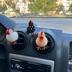 May include: Three felt chicken figurines, one black and white, one white, and one beige, are displayed in the air vents of a car's dashboard.