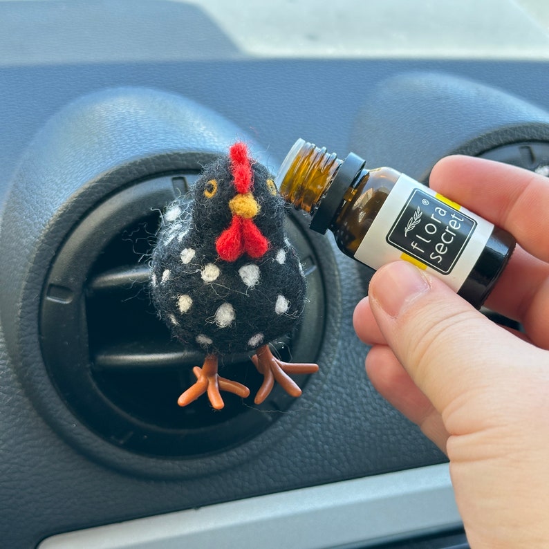 Puede incluir: Un clip de ventilaci&oacute;n de coche con forma de pollo negro y blanco con lunares, con una cresta roja y un pico amarillo. Se sostiene cerca una peque&ntilde;a botella de aceite esencial con el texto "flora secret".
