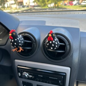 Puede incluir: Dos ambientadores con forma de pollo a lunares blancos y negros con crestas rojas y picos amarillos est&aacute;n sujetos a las rejillas de ventilaci&oacute;n del salpicadero de un coche. El salpicadero es gris y tiene una radio y controles de aire acondicionado.