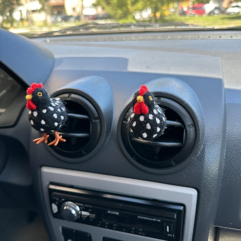 Puede incluir: Dos ambientadores con forma de pollo a lunares blancos y negros con crestas rojas y picos amarillos est&aacute;n sujetos a las rejillas de ventilaci&oacute;n del salpicadero de un coche. El salpicadero es gris y tiene una radio y controles de aire acondicionado.