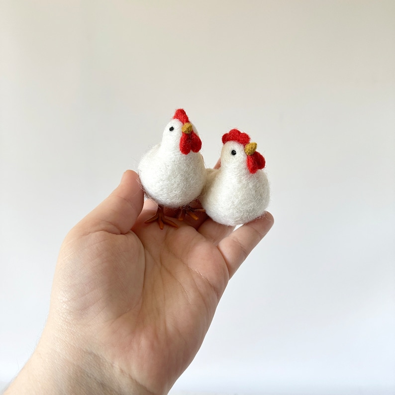 May include: Two white felt chickens with red combs and yellow beaks. The chickens are sitting in a person's hand.