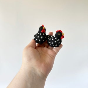 Puede incluir: Dos gallinas de fieltro hechas a mano. Las gallinas son negras con lunares blancos, crestas rojas y picos amarillos. Una gallina est&aacute; de pie y la otra est&aacute; sentada. Las gallinas son peque&ntilde;as y parecen ser objetos decorativos.